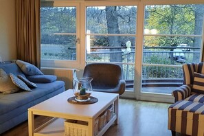 Ein Wohnzimmer mit Sofa, Sessel und Couchtisch, ein großes Fenster mit Blick auf Bäume und ein Balkon.