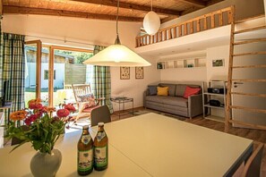 Ein gemütlicher Wohnbereich mit einer Couch, einem Tisch mit Blumen und Bierflaschen, einer Hängelampe und Blick durch eine Glastür.