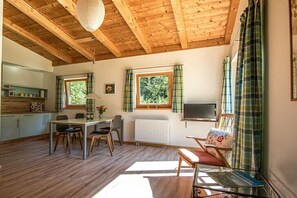 Ein Zimmer mit Holzboden, einem Esstisch mit Stühlen, einer Couch, einem Fernseher und einem Fenster mit Vorhängen.