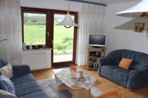 Ein Wohnzimmer mit einer Couch, einem Couchtisch mit Blumen, einem Fernseher auf einem Schrank und Blick durch eine Glastür.