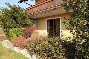A building with a balcony, a window with a white frame, and various shrubs and trees around it. - Quiet holiday apartment with lake view (Eggelsberg)