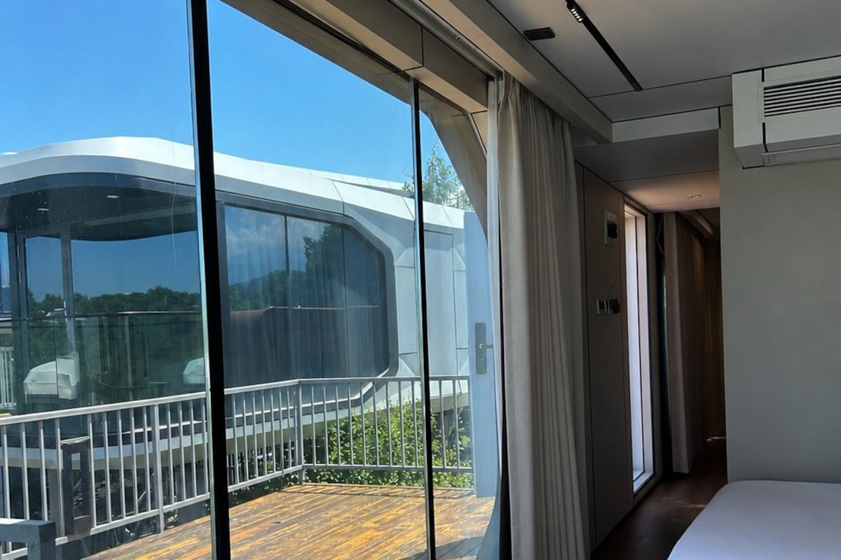 A modern hotel room with large windows, a balcony, and a bed.