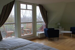Ein Zimmer mit einem Bett, zwei Stühlen, einem kleinen Tisch und Blick ins Freie durch große Fenster.
