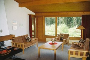 Ein gemütliches Wohnzimmer mit Holzmöbeln, einem Kamin und großen Fenstern mit Blick ins Freie.