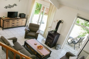 Ein Wohnzimmer mit Sofa, Sessel, Fernseher, Holzschrank, Kamin und Blick durch eine Glastür.