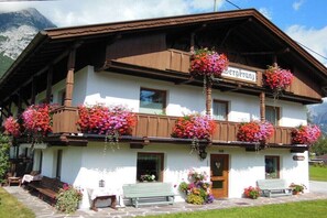 Ein traditionelles Chalet mit einem Balkon, geschmückt mit hängenden Blumenkörben, einem Kiesweg, der zum Eingang führt, und einem Bergpanorama im Hintergrund.