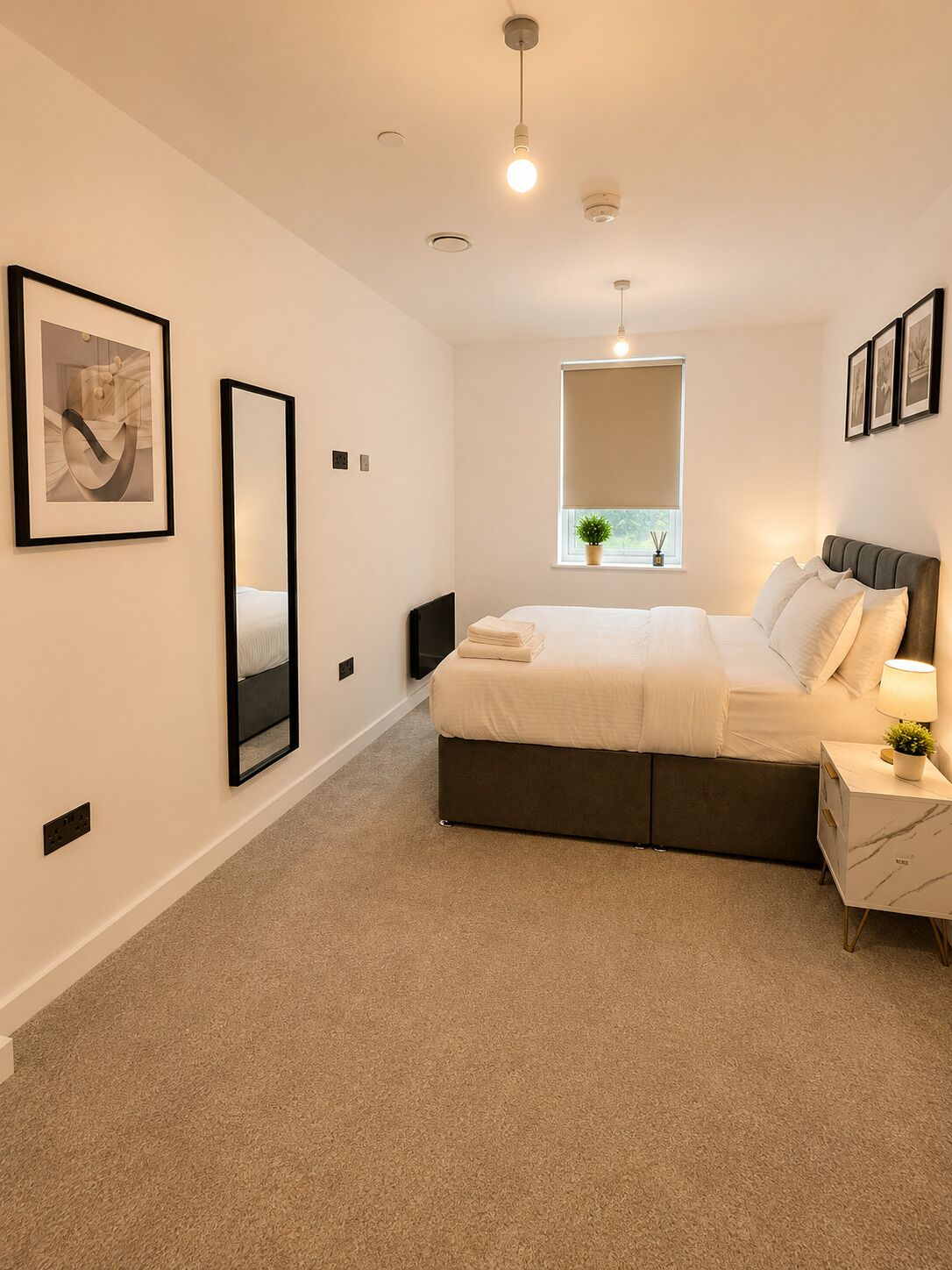 A bedroom with a bed, bedside table, lamp, window, and artwork on the wall.