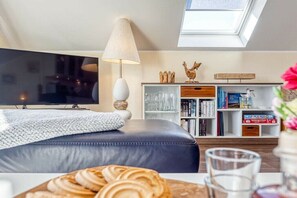 Ein modernes Wohnzimmer mit Flachbildfernseher, bequemer Couch, einem Esstisch mit Gebäckauslage, Bücherregal und einem Oberlicht.