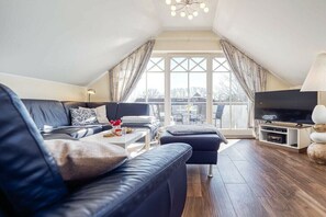 Ein modernes Wohnzimmer mit einer schwarzen Ledersofa, einem Couchtisch, einer TV-Konsole und einem großen Fenster mit Vorhängen.