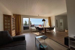 Ein Wohnzimmer mit einer Couch, einem Esstisch mit Stühlen, einem Glastisch und Blick ins Freie durch große Fenster.