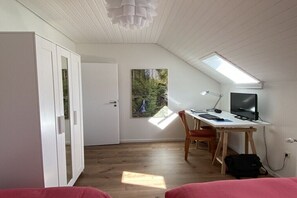 Ein Zimmer mit schräger Decke, einem Schreibtisch mit Computer, einem Stuhl, einem Fernseher und einem Fenster mit Blick auf Grünflächen.