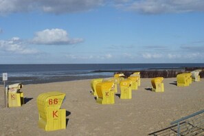 Ein Strand mit gelben Liegestühlen, die mit Zahlen und Buchstaben gekennzeichnet sind, einer Geländer und dem Meer im Hintergrund.
