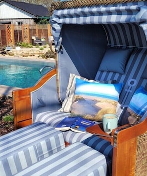 A striped lounge chair with a cushion, a small table with a book and a mug, and a pool in the background. - Holiday home Lüttes Paradies (Geestland)