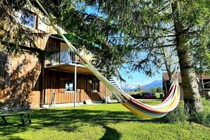 Eine Holzhütte mit Balkon, einer Hängematte und einem Picknicktisch in einer grünen Wiese mit Bäumen und Bergen im Hintergrund.
