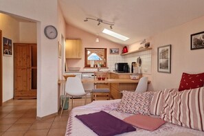 Ein kleines Schlafzimmer mit Bett, Schreibtisch und Stuhl, eine Küchenzeile mit Fenster sowie eine Uhr an der Wand.