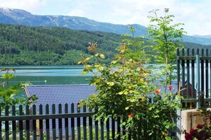 Blick auf einen See mit einer Holzwand, ein Gebäude mit Ziegeldach und einem Berg im Hintergrund.