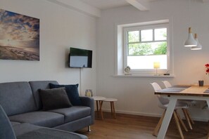 Ein modernes Wohnzimmer mit einem grauen Sofa, einem Esstisch mit Stühlen, einem an der Wand montierten Fernseher und einem Fenster mit Blick auf Grünflächen.