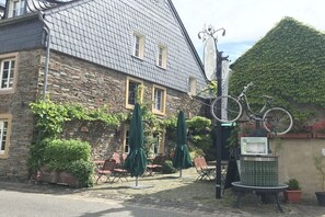 A stone building with a tiled roof, outdoor seating with umbrellas, and a bicycle mounted on a pole. - Holiday apartment with 1 bedroom (54347 Neumagen-Dhron, Deutschland)
