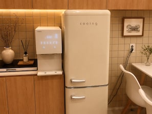 A kitchen with a white refrigerator labeled 'cooling,' a coffee machine, and a small table with a chair.