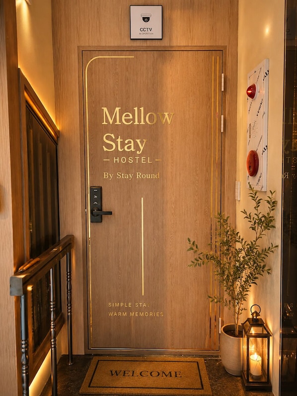 Una porta de fusta amb el rètol 'Mellow Stay Hostel', un senyal de CCTV, un estriau de 'BENVENUT' i una planta en test.