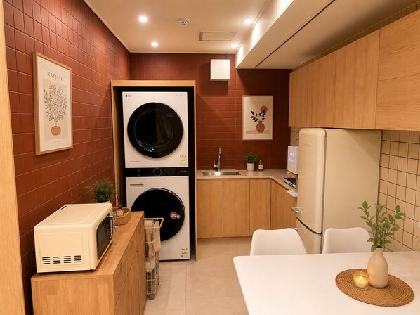 A compact kitchen with a washing machine, microwave, and refrigerator, wooden cabinets, and a small dining area.