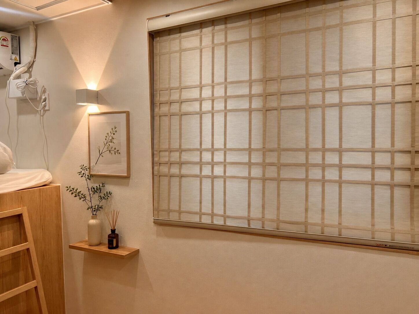 A room with a bed, a window with a grid pattern, a wall-mounted air conditioner, and a small shelf with a vase and reed diffuser.