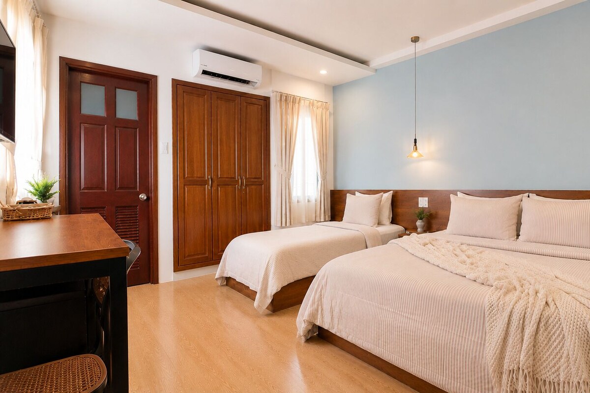 A bedroom with two beds, a wooden desk, a wardrobe, and an air conditioning unit.
