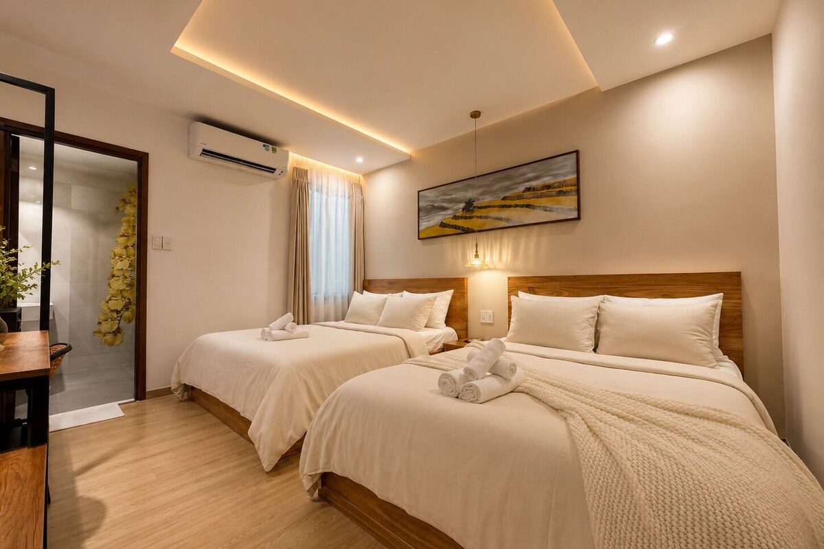 A hotel room with two beds, a wooden headboard, a painting, and a potted plant.
