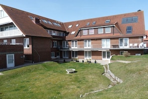 Ein zweistöckiges Backsteingebäude mit zahlreichen Fenstern, einer überdachten Veranda und einem Rasen mit Blumenbeeten.