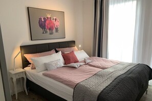 Ein ordentlich bezogenes Bett mit Kopfteil, ein Nachttisch mit Lampe und ein gerahmtes Bild an der Wand.