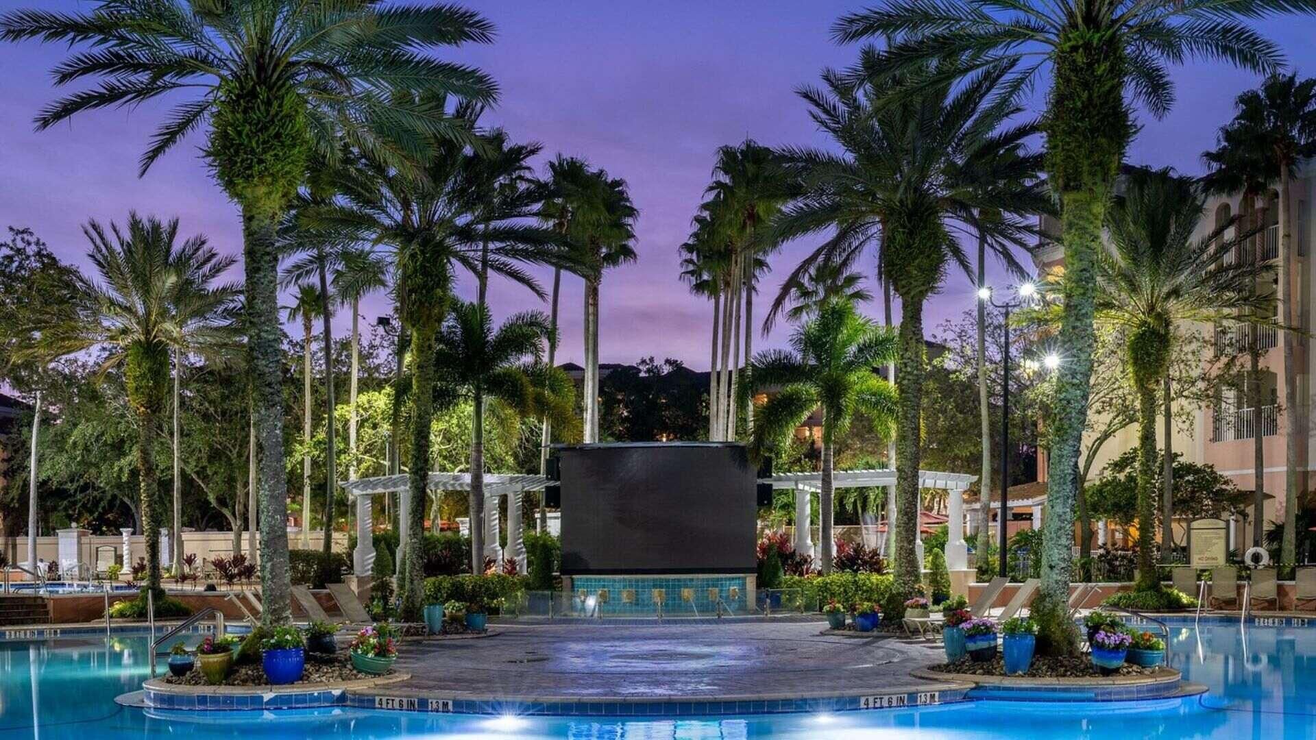 A swimming pool surrounded by palm trees and a covered area with a tiled floor.