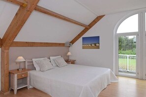Ein Schlafzimmer mit schrägverdeckter Decke, Holzbalken, einem großen Bett mit weißer Bettwäsche, zwei Nachttischen mit Lampen und Blick durch eine Glastür.