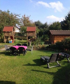 Un espace extérieur verdoyant avec une table et des chaises, deux cabanes en bois pour jouer et un petit chalet.