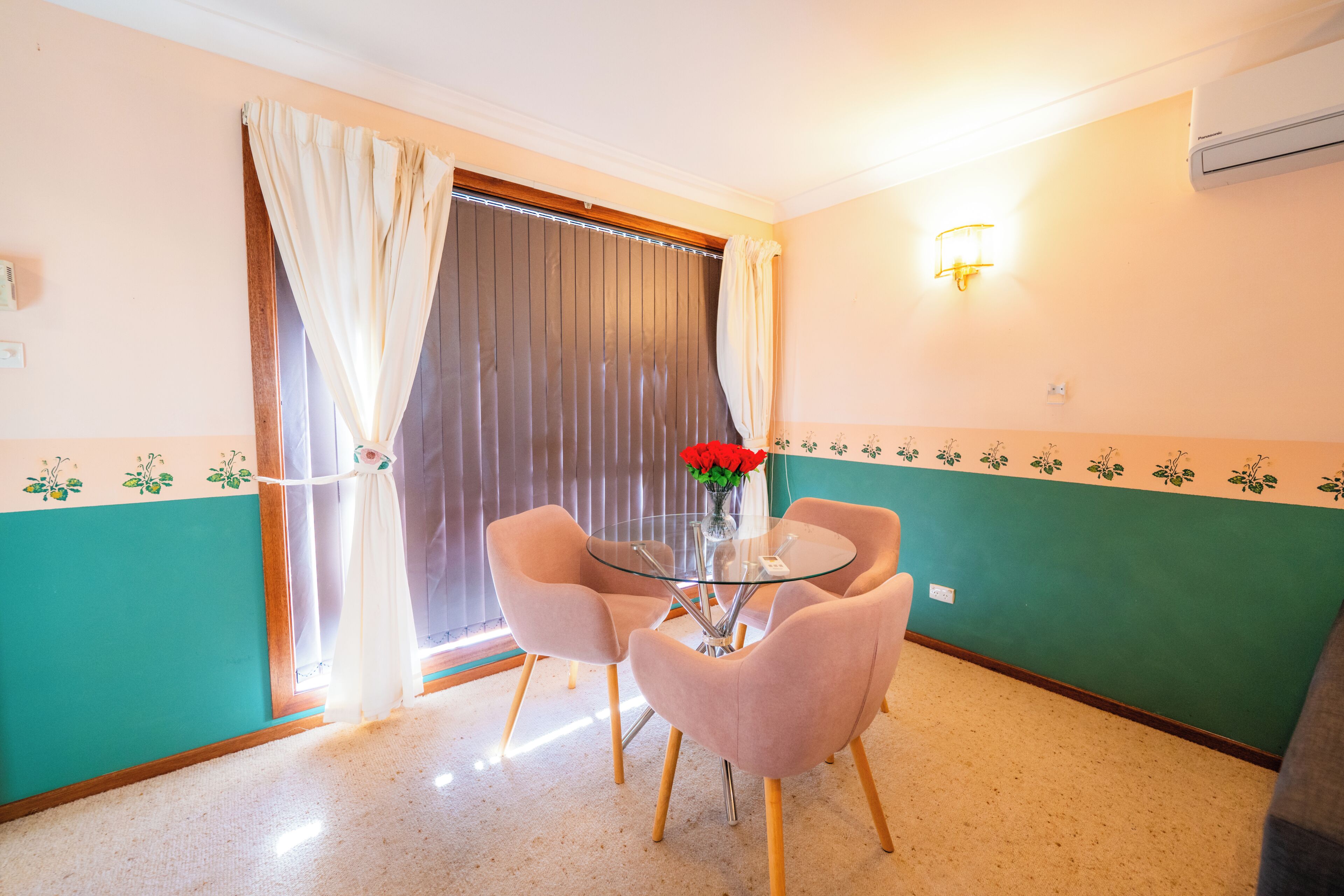 A room with a glass table, four chairs, a vase with flowers, a window with curtains, and a wall with a floral border.