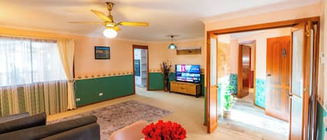 Una sala de estar con un sofá, una televisión, un ventilador de techo, un jarrón con flores y un pasillo que conduce a otra habitación.
