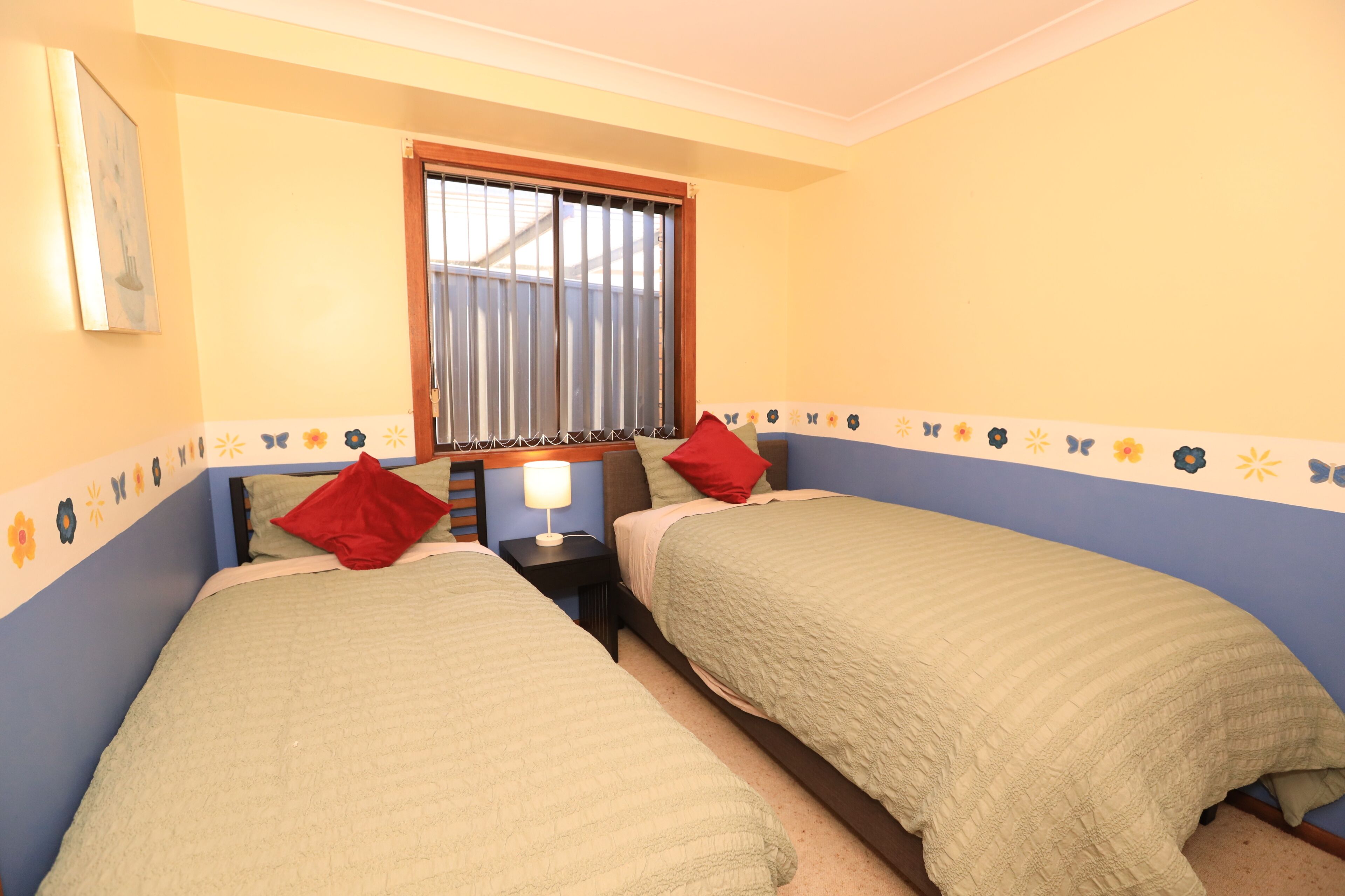 A bedroom with two beds, a window with blinds, and a decorative border on the walls.