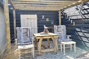 Ein rustikaler Sitzbereich im Freien mit einem Holztisch und -stühlen, einem Vogel auf dem Tisch und einer Holzhütte, auf deren Wand das Wort 'ZEE' steht.