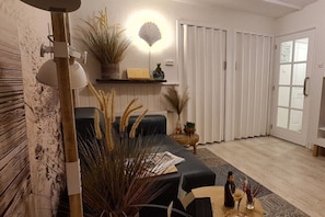 Ein modernes Wohnzimmer mit einer schwarzen Couch, Holzboden, einer weißen Wand mit einer leuchterartigen Lampe in Muschelform und einer Tür mit gefrostetem Glasfenster.
