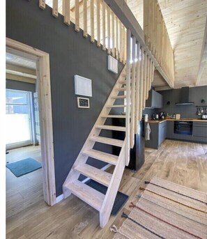 Eine Holztreppe führt zu einer zweiten Etage, ein Küchenbereich mit modernen Geräten und eine graue Wand mit einem kleinen Bilderrahmen.