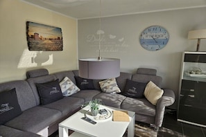 A living room with a grey sofa, a white coffee table, a clock, and a wall-mounted lamp. - Holiday apartment with 1 bedroom (Borkum)