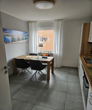 A small, well-lit room with a dining table, two chairs, a lamp, a window with curtains, and a kitchen area with a stove. - Holiday apartment with 1 bedroom (Borkum)