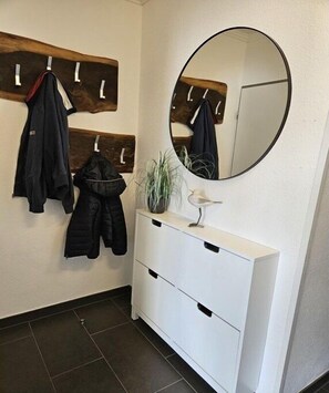 A bathroom with a wooden coat rack, a round mirror, a white cabinet with drawers, and a potted plant. - Holiday apartment with 1 bedroom (Borkum)