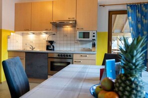 Eine kompakte Küche mit Mikrowelle, Herd und Spüle. Ein Esstisch mit Stühlen und einer Obstschale steht bereit.