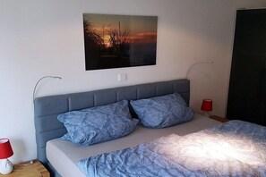 A bedroom with a bed, pillows, a nightstand, a lamp, a wall-mounted picture, and a door. - Schippers Huus No. 1 (Norderney)