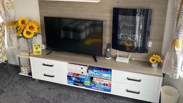 A TV stand with a flat-screen TV, a small shelf with decorative items, a vase with sunflowers, and a drawer with magazines. - Sunflower Lodge Luxury Pet Free Holiday Caravan (Filey)