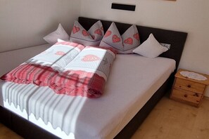 Ein ordentlich bezogenes Bett mit einer weiß-rot karierten Decke, einem dunklen Kopfteil und einem Nachttisch aus Holz mit einer Spitzendecke.
