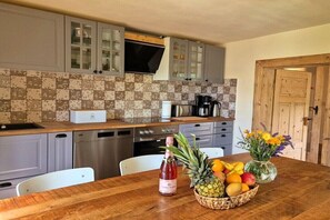 Eine Küche mit grauen Schränken, einer Holz Arbeitsplatte, einer Kaffeemaschine, einem Geschirrspüler und einem Esstisch mit einer Champagnerflasche, einer Obstschale und einem Blumenstrauß.