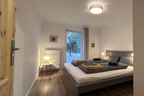Ein Schlafzimmer mit Bett, Nachttischlampen, einem Heizkörper, einem Fenster mit Vorhängen und Blick ins Freie.