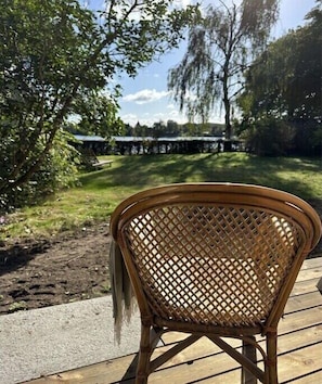 Ein Korbstuhl auf einer Holzterrasse mit Blick auf einen Garten und ein Gebäude im Hintergrund.