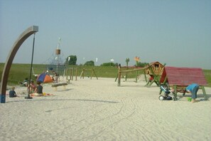 A sandy beach with a swing set, a small hut, and a fenced area. - Hansmeier in the Eckwarderhörne holiday village (Butjadingen)